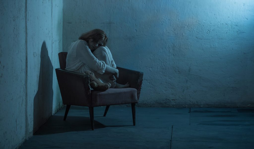 Copil singur, pe fotoliu, într-o cameră întunecată, cu jucăria de pluș lângă el