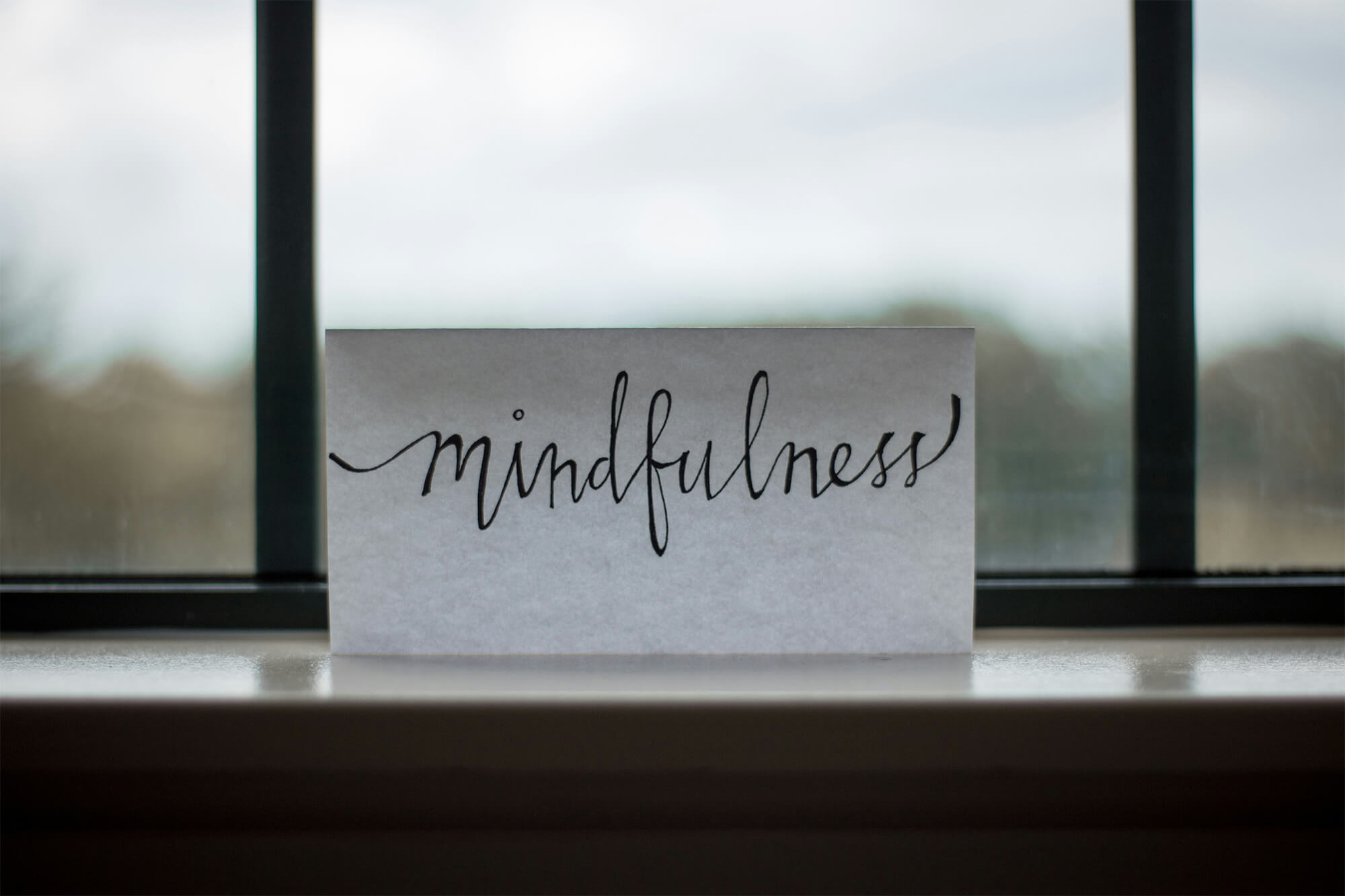 Un cartonaș cu inscripția „mindfulness” scrisă elegant, așezat pe pervazul unei ferestre, cu fundal estompat.