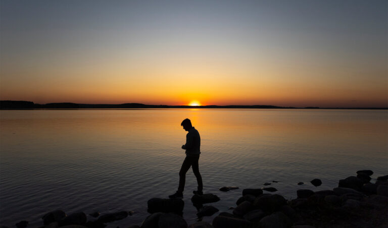 Silueta unei persoane care merge pe pietre, la marginea unui lac liniștit, gândindu-se la gândurile sinucigașe pe care le are, în timpul unui apus de soare cu nuanțe calde de portocaliu și albastru pe cer și pe apă. Atmosfera este liniștită și contemplativă.