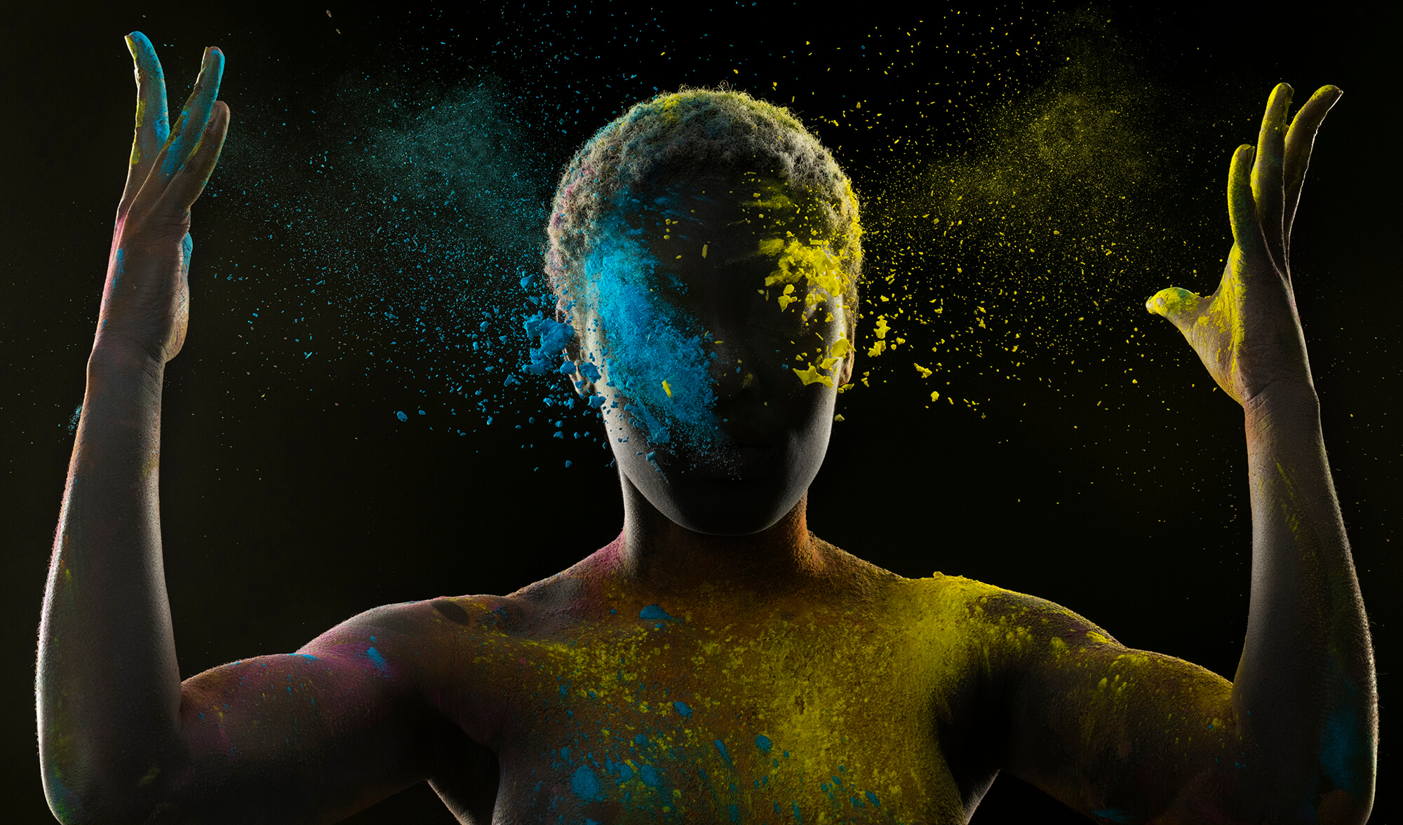 O siluetă umană cu brațele ridicate în semn de victorie, acoperită cu pudră colorată în albastru și galben care explodează dinamic în jurul capului, simbolizând optimismul în creier ca o forță vibrantă și transformatoare care radiază energie pozitivă în toate direcțiile.